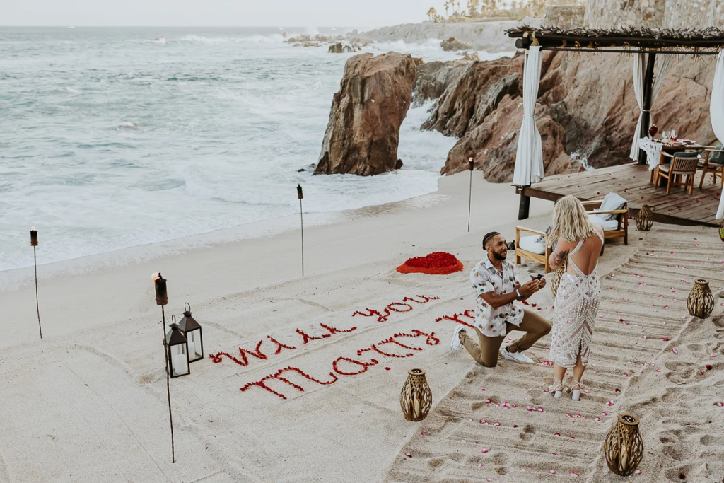 man on his knee proposes to future bride, you can read "will you marry me" on the sand with flower petals