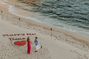 Surprise proposal at Esperanza Los Cabos, man is on one knee and you can read "Marry Me" on the sand