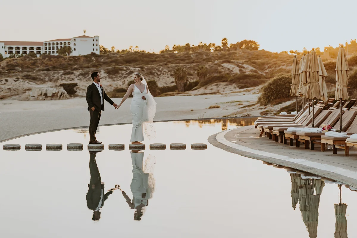 Bride and groom walking, she is leading him and their reflection is shown in water