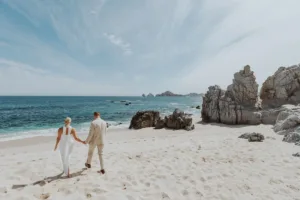 Bride and groom walk at the beach holding hands, the famous Cabo landmark with the arch is in the background